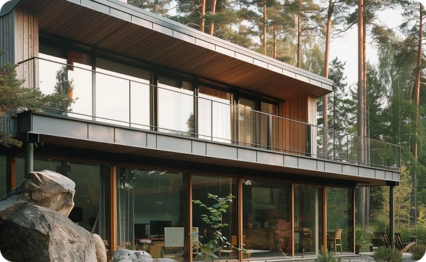 Dit is een moderne woning met twee verdiepingen, gelegen in een uitgestrekt bosrijk gebied, omringd door hoge bomen. De buitenomgeving straalt gezelligheid en elegantie uit, met natuursteen en kleine struiken verspreid over het terrein voor het huis. De stijl van de foto is die van een professionele opname uit een fotoshoot of architectuurreportage, waarbij de nadruk ligt op de harmonie tussen het huis en de natuur en de strakke lijnen van de architectuur. Het kleurenpalet bestaat voornamelijk uit tinten van natuurlijke materialen: grijs, bruin, groen en warm zonlicht, wat een gezellige en aangename sfeer creëert.