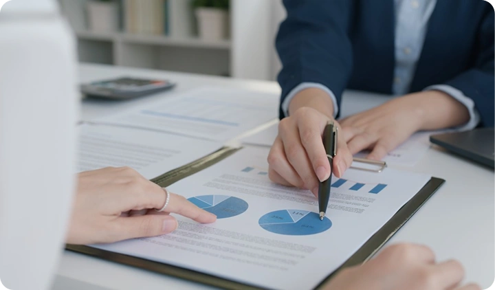 In het middelpunt staan de handen van twee personen: de ene houdt een pen vast en wijst naar een grafiek met cirkeldiagrammen en tekstblokken op een vel papier, de andere wijst met zijn vinger naar het diagram. Beide personen dragen zakelijke kleding: de ene een pak met een lichtgekleurd colbert en een overhemd, de andere een donker colbert en een licht overhemd. Op de tafel liggen een laptop, enkele documenten en een map met papieren. De sfeer is rustig en zakelijk; de foto is professioneel, in close-up genomen, met de focus op de handen en het document. Het kleurpalet van de foto bestaat uit zachte pasteltinten, waarbij blauw, wit en lichte tinten overheersen.